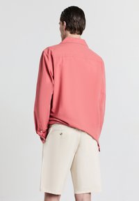 Chemise à manches longues de couleur corail avec un col boutonné, associée à un short de couleur crème. La chemise présente une texture lisse et une coupe décontractée.