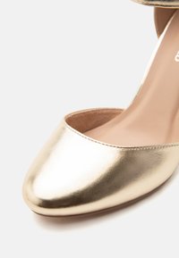 Λαμπερό χρυσό γυναικείο παπούτσι στιλέτο με κλειστή μύτη, στρογγυλεμένο μπροστινό μέρος και λείο δερμάτινο φινίρισμα σε λευκό φόντο.