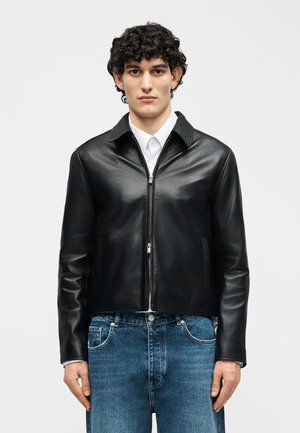 Jeune homme aux cheveux bouclés portant une veste en cuir noire zippée par-dessus une chemise blanche et un jean bleu, debout devant un fond uni.