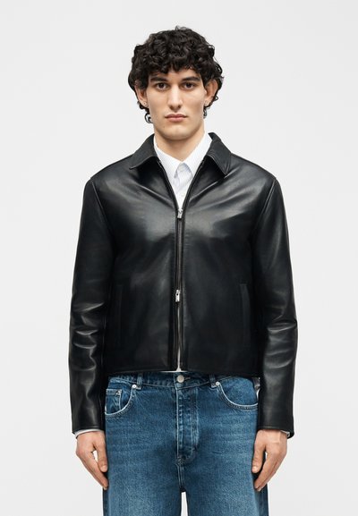 Jeune homme aux cheveux bouclés portant une veste en cuir noire zippée par-dessus une chemise blanche et un jean bleu, debout devant un fond uni.