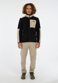 Hombre de pie de frente, vistiendo una chaqueta de forro polar negra y beige, pantalones beiges, y zapatos de senderismo verdes y negros sobre un fondo blanco.