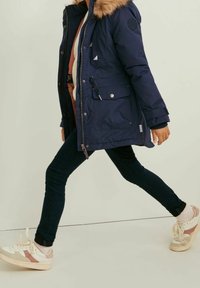 Navy parka with faux fur hood, zip closure, and pockets. Paired with dark jeans and pastel sneakers featuring a color-block design.
