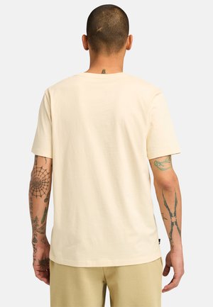 Cremefarvet, kortærmet t-shirt lavet af bomuld, med et lige snit. Bagfra ses tatoveringer og en ren halsudskæring uden logoer.