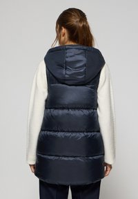 Gilet en duvet marine avec capuche, doté de panneaux mats horizontaux et d'un col haut, porté par-dessus un pull de couleur claire. Texture douce.