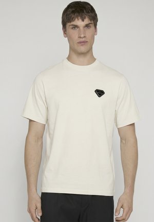 Mand iført en ensfarvet hvid T-shirt med korte ærmer og et lille sort diamantformet logo på venstre bryst, stående foran en ensfarvet baggrund.
