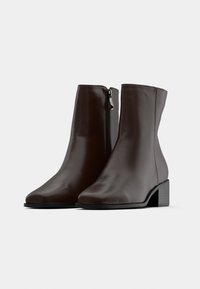 Paire de bottines en cuir marron foncé avec fermeture éclair latérale et petits talons carrés sur un fond gris.