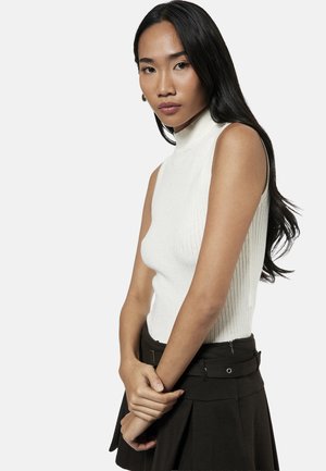 Junge Frau mit langen schwarzen Haaren, die ein weißes ärmelloses Rollkragentop und einen schwarzen Rock trägt und mit vor dem Körper verschränkten Händen posiert.