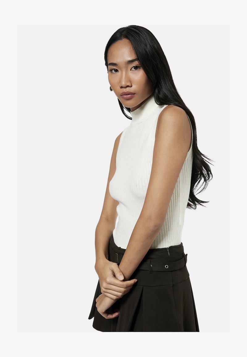 Junge Frau mit langen schwarzen Haaren, die ein weißes ärmelloses Rollkragentop und einen schwarzen Rock trägt und mit vor dem Körper verschränkten Händen posiert.