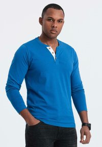 Camicia henley a maniche lunghe blu con pattina bianca e dettagli marroni. Abbinata a jeans scuri, caratterizzati da una superficie liscia e un design aderente.