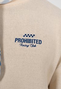 Pull en tricot beige clair avec tissu texturé. Présente le texte brodé bleu "PROHIBITED" et "Racing Club" avec un motif à damier au-dessus.