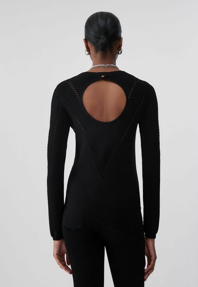 Femme aux cheveux noirs en chignon porte un pull noir à manches longues avec une grande découpe ovale dans le haut du dos, assorti d'un pantalon noir.