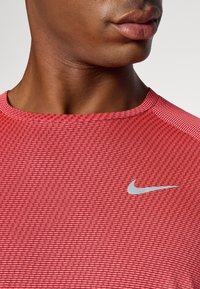 Piros sporting ing, textúrált anyagból készült. Kerek nyakkivágással rendelkezik, és egy kis ezüst Nike logó található a bal mellen.