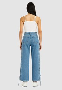 Jeans a vita alta azzurri chiari con gambe larghe, due tasche posteriori e lacci sui lati inferiori, abbinati a una canotta bianca e sneakers bianche.