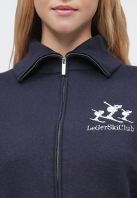 Tumši zils fleeces džemperis ar rāvējslēdzēju un augstu apkakli, ar baltu slēpotāju izšuvumu. Izstrādāts siltumam, ar gludu tekstūru un smalku spīdumu.