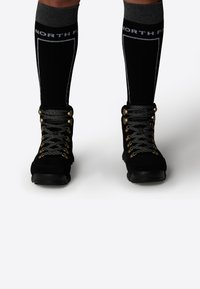 Bottes de randonnée noires en daim avec des lacets gris et des accessoires dorés. Associées à des chaussettes noires jusqu'au genou avec un bandeau supérieur gris et un logo.