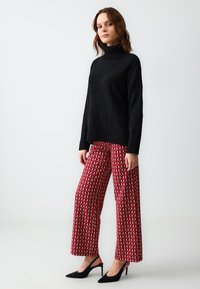 Maglione a collo alto nero in maglia, abbinato a pantaloni larghi rossi con una fantasia geometrica, completato da scarpe con tacco nere.