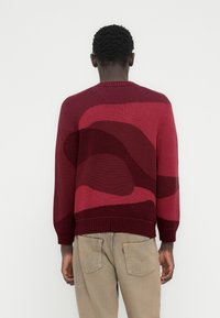 Μπορντό πουλόβερ με αφηρημένα πιο σκούρα μπορντό σχέδια, στρογγυλή λαιμόκοψη, ριμπ μανσέτες και τελείωμα. Φορεμένο πάνω από ανοιχτό μπεζ παντελόνι.