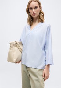 Νεαρή γυναίκα με κοντά ξανθά μαλλιά, φοράει ανοιχτό μπλε μπλούζα με V λαιμόκοψη και μπεζ παντελόνι, κρατώντας μια μεγάλη μπεζ τσάντα στο αριστερό της χέρι.