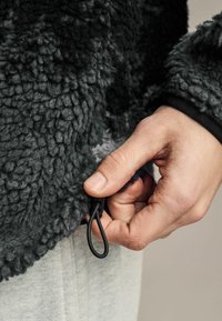 Ciemnoszara, pluszowa kurtka z polaru o teksturowanej powierzchni. Ręka ciągnie za czarną sznurówkę na dole, demonstrując funkcję regulacji.