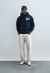 Man wearing black graphic hoodie, off-white loose pants, white sneakers, standing with hands in pockets against plain light gray background.