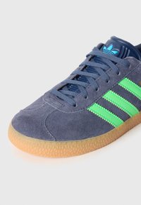 Modre antilop superge z zelenimi črtami, teksturirano gumijastim podplatom in okrolimi prsti. Ponašajo se z modrimi vezalkami in temno modrim oblazinjenim ovratnikom.