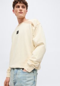 Νέος άνδρας με σγουρά μαλλιά που φοράει μπεζ φούτερ με λεπτομέρεια πατς και ανοιχτόχρωμο μπλε τζιν, με τα χέρια στις τσέπες, κοιτάζοντας προς τα αριστερά.