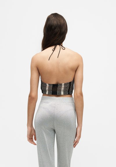 Femme aux longs cheveux bruns portant un crop top dos nu noir et blanc et un pantalon argenté taille haute, debout devant un fond clair uni.