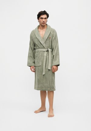 Homme debout pieds nus portant un peignoir en éponge vert clair noué à la taille avec deux poches avant et un petit logo rouge sur la poitrine.