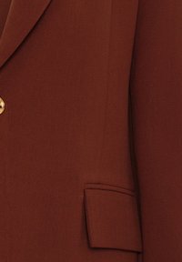 Gros plan sur une veste marron avec un bouton doré et une poche à rabat avant, montrant la texture du tissu et les coutures ajustées.
