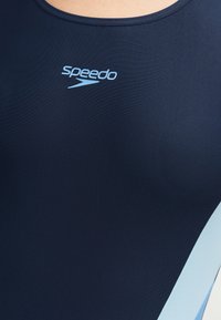 Μαγιό Speedo σε ναυτικό μπλε χρώμα με ανοιχτό μπλε πλαϊνά πάνελ, φορεμένο από άτομο, με εμφανές το άνω μέρος του στήθους και το λογότυπο στο κέντρο κάτω από τον λαιμό.