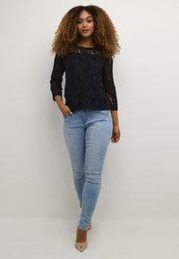 Black lace top with ¾ sleeves, featuring a scalloped neckline and floral pattern, paired with light blue fitted jeans and beige heels.