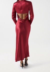 Czerwona satynowa sukienka z długimi rękawami, otwartym plecami i marszczonym wystrojem. Długość do podłogi, o gładkiej fakturze i minimalistycznej sylwetce. Czarne szpilki.
