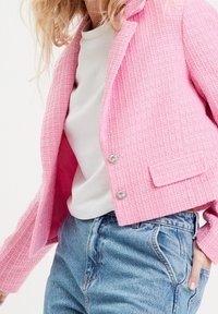 Blazer rose texturé avec un motif tissé, des revers crantés et des boutons argentés, associé à un haut blanc et un jean en denim taille haute.