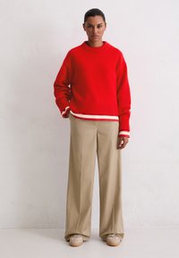 Czerwony sweter z dzianiny z prążkowanym kołnierzem i mankietami, noszony na białym paskowanym dołem, w połączeniu z beżowymi spodniami o szerokich nogawkach i beżowymi trampkami.