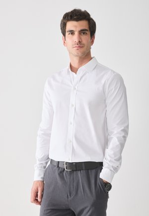 Camisa blanca de algodón de manga larga con botones y cuello, combinada con pantalones grises y un cinturón negro, luciendo un reloj en la muñeca izquierda.