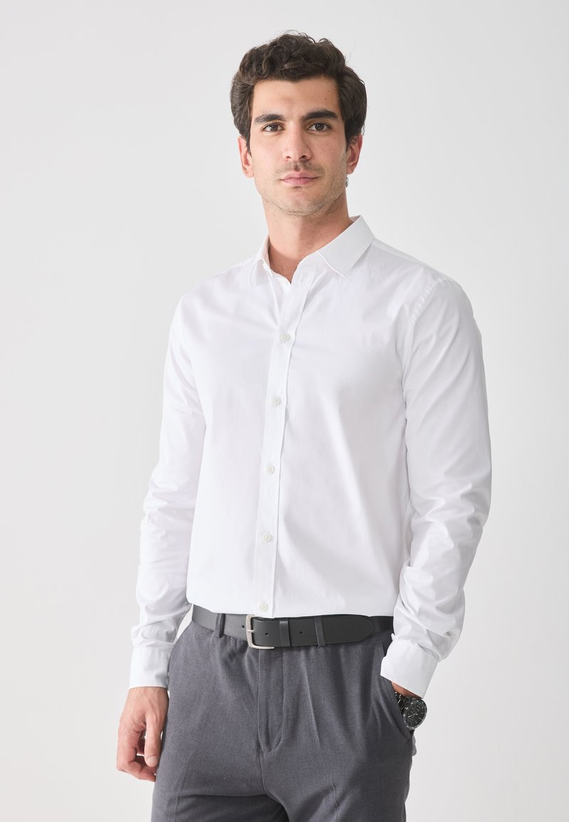White cotton long-sleeve button-up shirt with a collar, paired with gray trousers and a black belt, featuring a watch on the left wrist.
