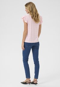 Lys pink top med flæser på ærmerne og hulmønsterdetaljer, parret med blå skinny jeans og sorte spidse flade sko. Bagfra vist.