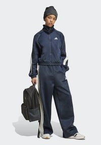 Navy blue adidas tracksuit with white side stripes, zip-up jacket, wide-leg pants, and a beanie; model holds a black backpack.