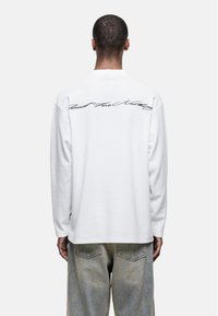 Long-sleeve, white cotton shirt featuring ribbed texture and cursive text on the back. Paired with light-colored denim jeans.