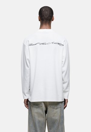 Chemise blanche en coton à manches longues avec texture côtelée et texte en cursive au dos. Associée à un jean en denim de couleur claire.