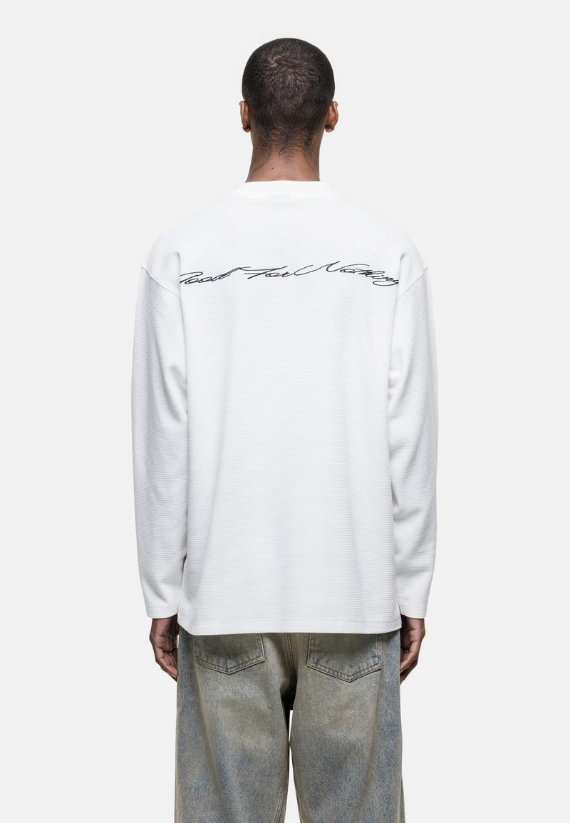 Long-sleeve, white cotton shirt featuring ribbed texture and cursive text on the back. Paired with light-colored denim jeans.