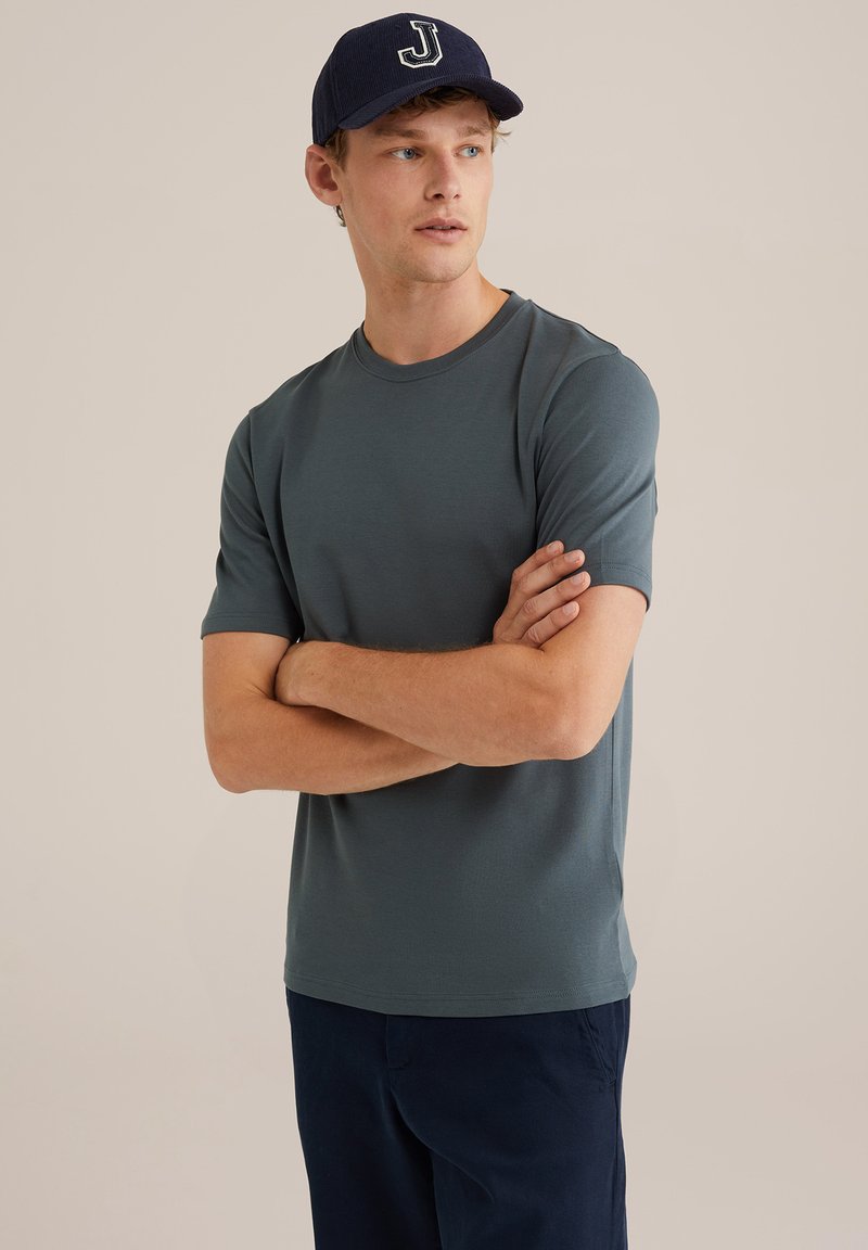 Camiseta de manga corta para hombre, de cuello redondo, en verde azulado, hecha de una tela suave. Lleva puesta una gorra azul marino con el logotipo blanco de la 'J'; brazos cruzados.