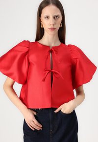 Top corto in satin rosso con maniche a sbuffo, dotato di una apertura a chiave legata con due fiocchi sul collo. Texture liscia e design strutturato.
