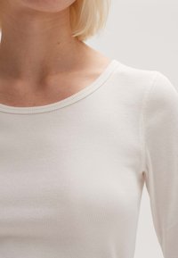 Långärmad vit ribbad topp med rundad halsringning och mjuk textur, med subtila sömdetaljer längs axlarna.