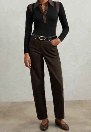 Person trägt ein schwarzes Langarmshirt mit braunem Kragen und Einsätzen, dunkelbraune Hose mit schwarzem Gürtel und braune Ballerinas, steht auf Betonboden.