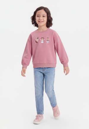 Lille barn med krøllet hår iført en lyserød sweatshirt med ballerina-prints, lyseblå jeans og lyserøde sneakers, som går og smiler.