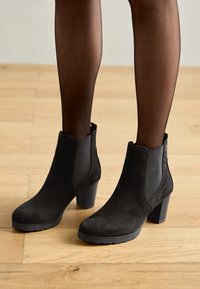 Svarta mockaskor anklar med elastiska sidopaneler och blockklackar. Texturerad yta och rund tå, placerad mot ett trägolv.