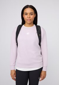 Ung kvinna med långt mörkt hår som bär en ljuslila sweatshirt, svart ryggsäck och svarta byxor, vänd mot kameran framför en neutral bakgrund.