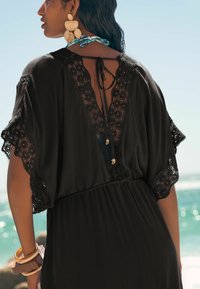 Robe noire ample avec un dos en dentelle en V, des manches en dentelle festonnées et un lien de fermeture au niveau du cou. Accentuée de détails dorés.
