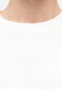 Κοντινό πλάνο στον λαιμό και την κλείδα ενός ατόμου που φοράει ένα απλό λευκό πουκάμισο με στρογγυλή λαιμόκοψη, πάνω σε λευκό φόντο.